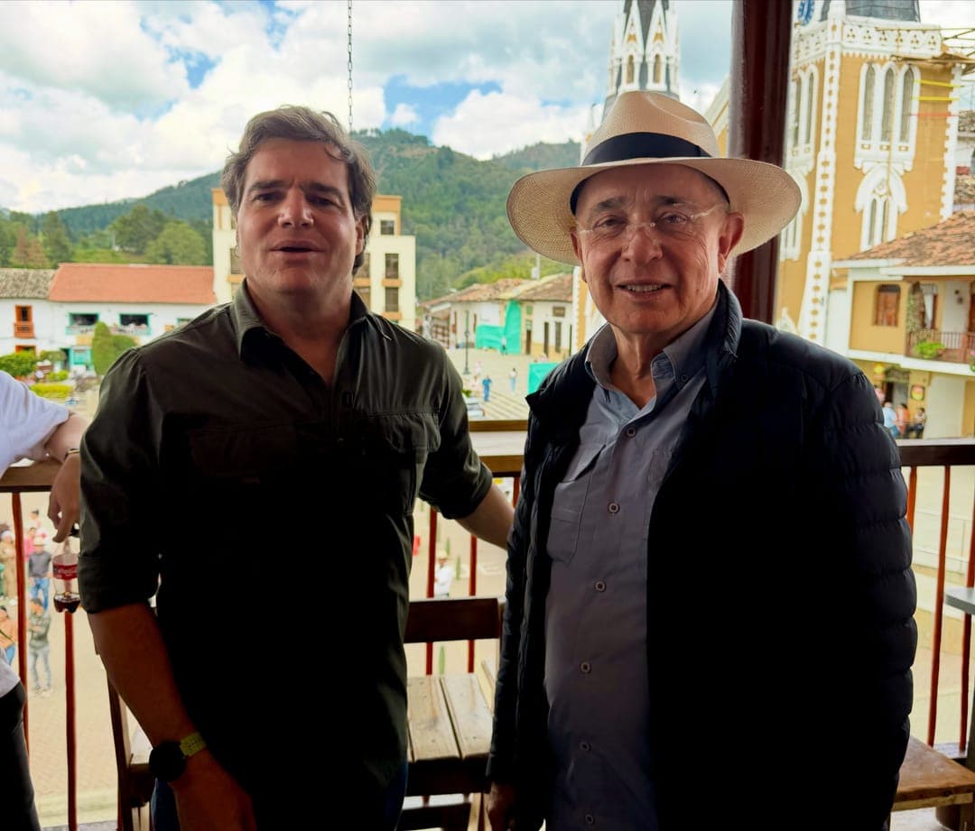 Alejandro Arbeláez A. y Álvaro Uribe Vélez, con sombrero, en un balcón frente a la plaza y la iglesia de un pueblo antioqueño